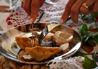 Na kolorowym talerzu leżą dwa opiekane pierogi i porcja kremowego sosu. Talerz stoi na stole nakrytym białą koronkowa serwetą. Widać kobiece dłonie, które widelcem i nożem kroją potrawę.