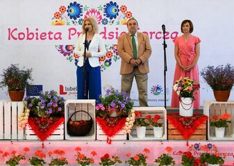 Trzy osoby stoją na scenie ozdobionej kwiatami. Zanimi widoczna jest biała ścianka z dużym sercem z kwiatów w motywie folkowym i napisem Kobieta Przedsiębiorcza