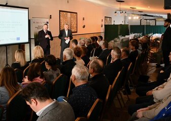 Grupa osób siedząca na krzesłach i słuchająca przemowy mężczyzny ubranego elegancko