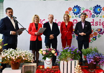 Pięć osób stoi na scenie. Wśród nich Pan Zdzisław Szwed, Członek Zarządu Województwa Lubelskiego, Pani Ewa Szałachwiej, Dyrektor Departamentu Rolnictwa i Rozwoju Obszarów Wiejskich w Urzędzie Marszałkowskim w Lublinie i Pan Henryk Karwan, Starosta Powiatu Tomaszowskiego stoją na scenie udekorowanej kwiatami. W tle widać białą ściankę z dużym sercem z kolorowych kwiatów w motywie folkowym i napisem "Kobieta Przedsiębiorcza"