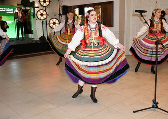 Trzy kobiety, śpiewają, ubrane w kolorowe stroje ludowe. W tle scena , na której siedzi mężczyzna ubrany w strój ludowy, gra na akordeonie