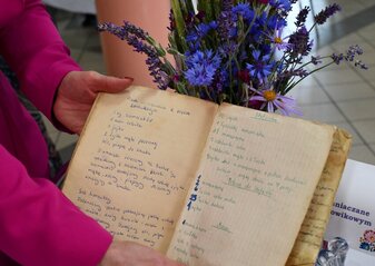 Stary pożółkły zeszyt z pisanym odręcznie przepisem kulinarnym trzymany7 w rękach kobiety. W tle widać bukiet kwiatów