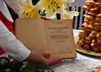 Stara ksiażka kucharska 1933 roku trzymana w dłoniach kobiety na tle kwiatów i ciasta sękacz