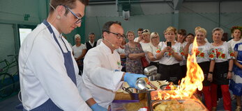 Pokaz kulinarny, dwóch kucharzy przy stole, wokół stołu stoją kobiety. Na stole produkty spożywcze, naczynia oraz urządzeni kuchenne. Jedno z naczyń jest palnikiem podpalane przez kucharza.