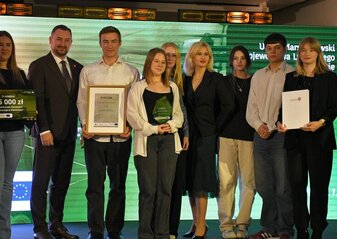 Na scenie, na tle kolorowej grafiki stoi grupa osób (pięć kobiet i trzech mężczyzn) ubranych elegancko, w dłoniach trzymają czek, statuetkę, dyplom i białą teczkę