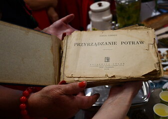 Książka kulinarna z 1936 roku trzymana w dłoniach kobiety. Książka ma pożółkłe kartki i luźne strony, jest bardzo stara.