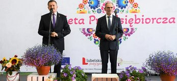 Na scenie stoi Pan Zdzisław Szwed, Członek Zarządu Województwa Lubelskiego i Pan Zdzisław Antoń, Starosta Lubelski. Za nimi widoczna jest biała ścianka z dużym sercem z kwiatów w motywie folkowym z napisem przez środek "Kobieta Przedsiębiorcza"