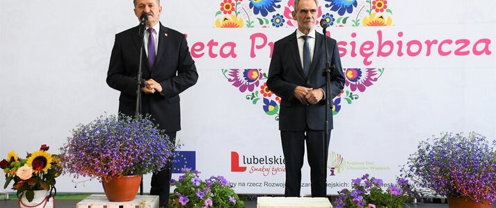 Na scenie stoi Pan Zdzisław Szwed, Członek Zarządu Województwa Lubelskiego i Pan Zdzisław Antoń, Starosta Lubelski. Za nimi widoczna jest biała ścianka z dużym sercem z kwiatów w motywie folkowym z napisem przez środek "Kobieta Przedsiębiorcza"