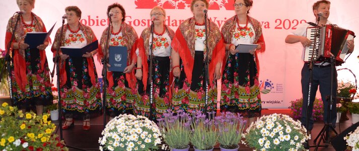 Ludowy zespól muzyczny. 6 kobiet ubranych w stylu ludowym, z czerwonymi chustami na ramionach, po prawej stronie mężczyzna z akordeonem. Przed sceną dekoracja z różnokolorowych kwiatów. W tle czerwone serce z wzorów łowickich, przez środek, którego na białym pasku przechodzi napis Kobieta Gospodarna Wyjątkowa 2022