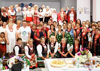 Duza grupa osób stoi przed sceną udekorowaną kwiatami. Są to uczestnicy półfinału konkursu Kobieta Przedsiębiorcza. Uczestnicy półfinału konkursu ubrani są w regionalne stroje ludowe