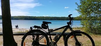 Rower oparty o ławkę stoi na koło drzewa. W tle widać jezioro.