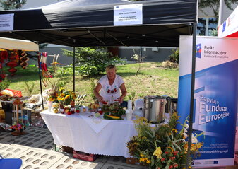 Na zdjęciu widoczne stoisko Koła Gospodyń Wiejskich prowadzające pokaz kulinarny wraz z degustacją zupy pokrzywowej. Na stole ustawione ozdoby z ziół, produkty spożywcze. Po lewej stronie stoją duże termosy z zupa pokrzywową 