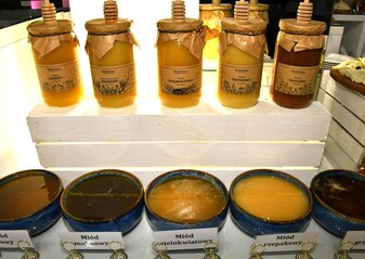 Na półce stoi pięć słoików z miodem. Jest to miód, kolejno od lewej: lipowy, malinowy, wielokwiatowy, rzepakowy i gryczany. Przed słoikami stoją miseczki z miodem do degustacji